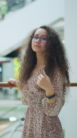 mujer en vestido vintage posa pensativamente coloca la mano en la cintura cerca de la barandilla de hierro, ajustando su cabello en un centro comercial brillante, el borrón de fondo destaca a la gente en la escalera mecánica en movimiento y la planta en maceta cercana