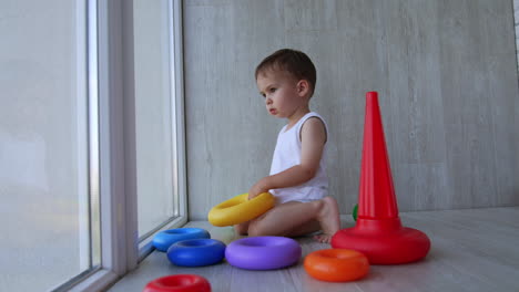 cute baby boy in white bodysuit sits on the floor holding a toy. serious kid looks into the window and says something.
