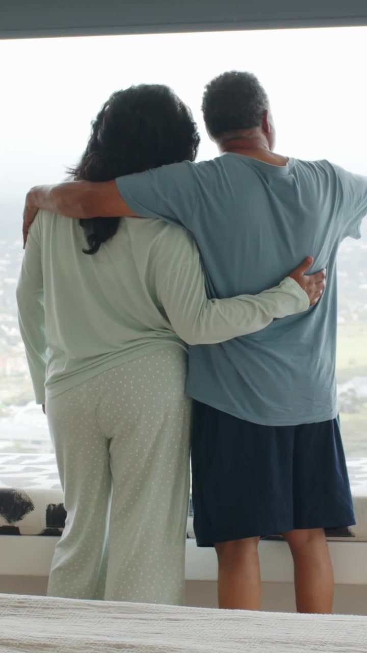 Video verticale: Coppia che si abbraccia davanti a una grande finestra, guardando fuori, con abiti verdi e maglietta blu.