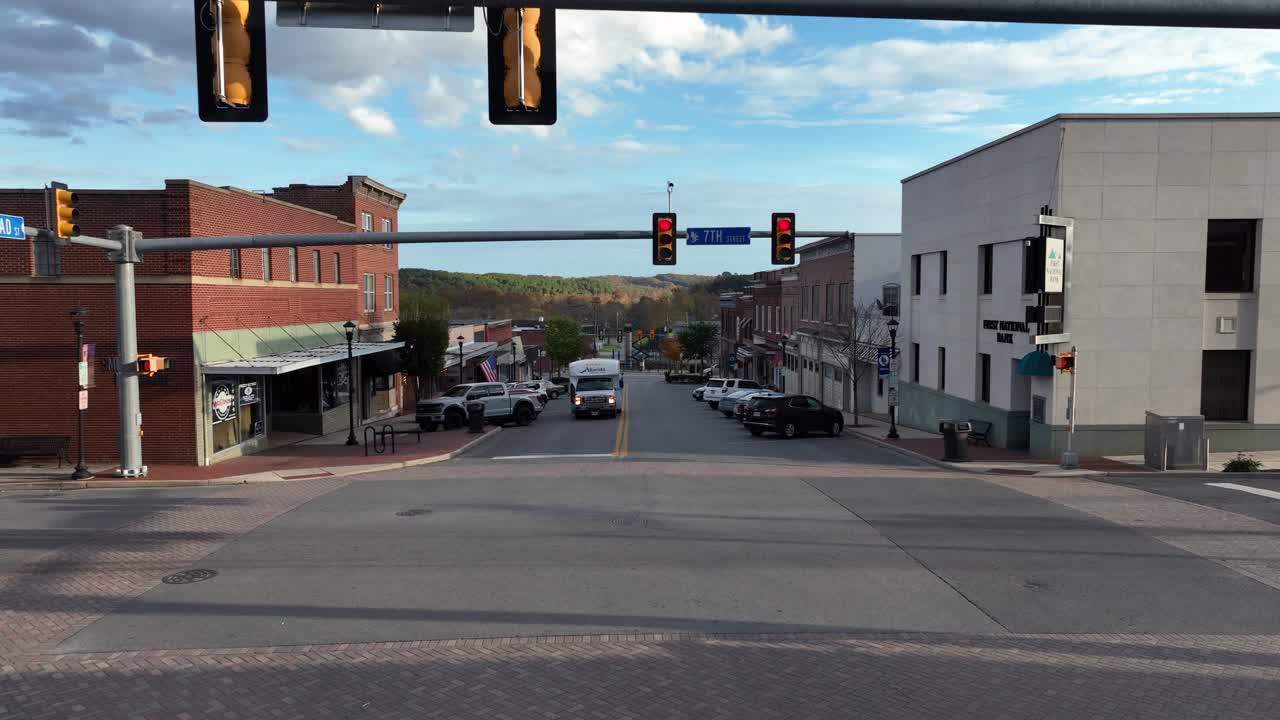 Aerial flight over junction in american town during red lights. Altavista City Bus and parking cars along street. First national bank and barber shop with american flag at sunset. Wide shot.