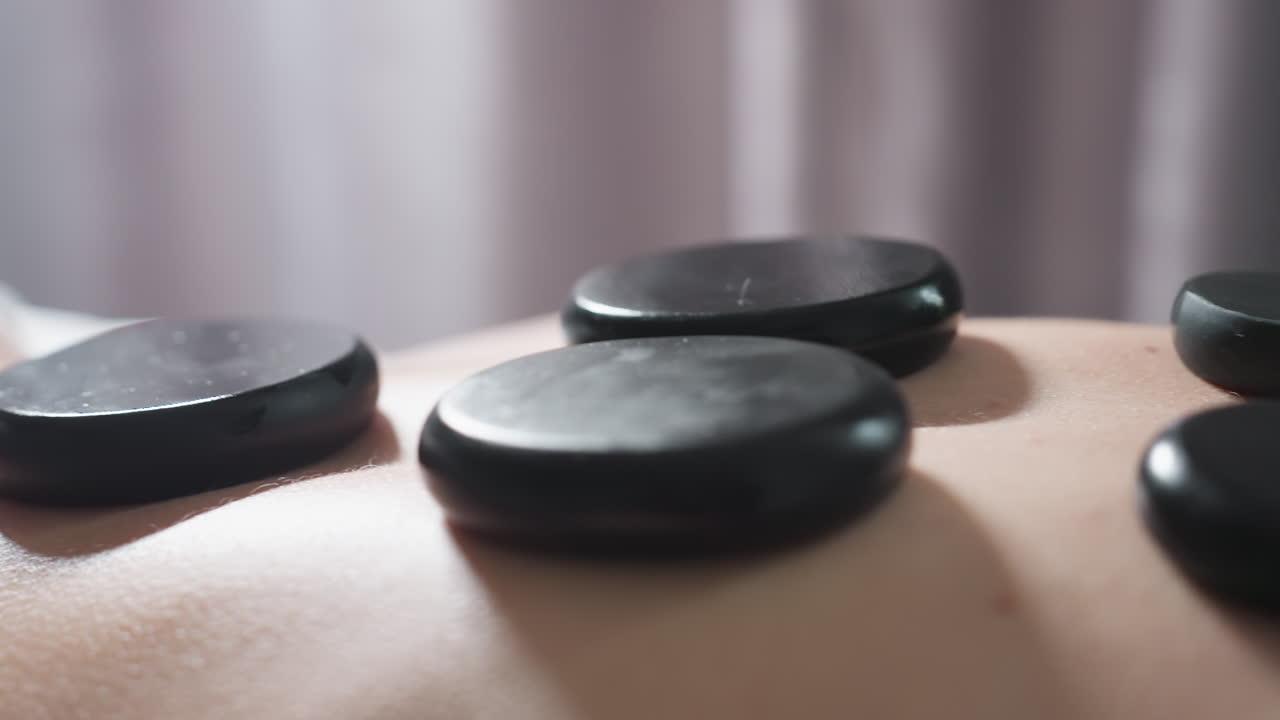 Very close up view of smooth massage stones arranged on client back during spa therapy session with soft light and blurred curtain background, capturing warmth, texture, and relaxation atmosphere