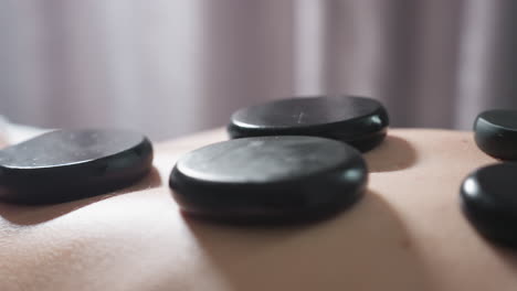 very close up view of smooth massage stones arranged on client back during spa therapy session with soft light and blurred curtain background, capturing warmth, texture, and relaxation atmosphere