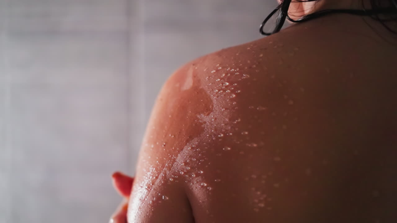 Woman applies scrub on shoulder in shower unit closeup. Lady exfoliates dry skin with homemade cosmetic product in bathroom. Spa relaxation at home