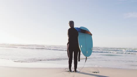 Ältere-Afroamerikanische-Frau,-Die-Mit-Einem-Surfbrett-Am-Strand-Spaziert