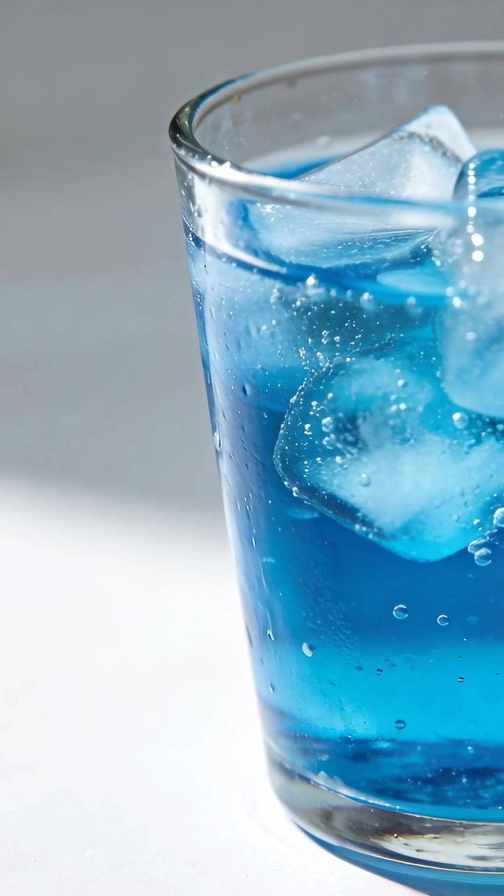 Vertical video: Panning glass showing blue soda with ice on white table to show bubbles, copy space