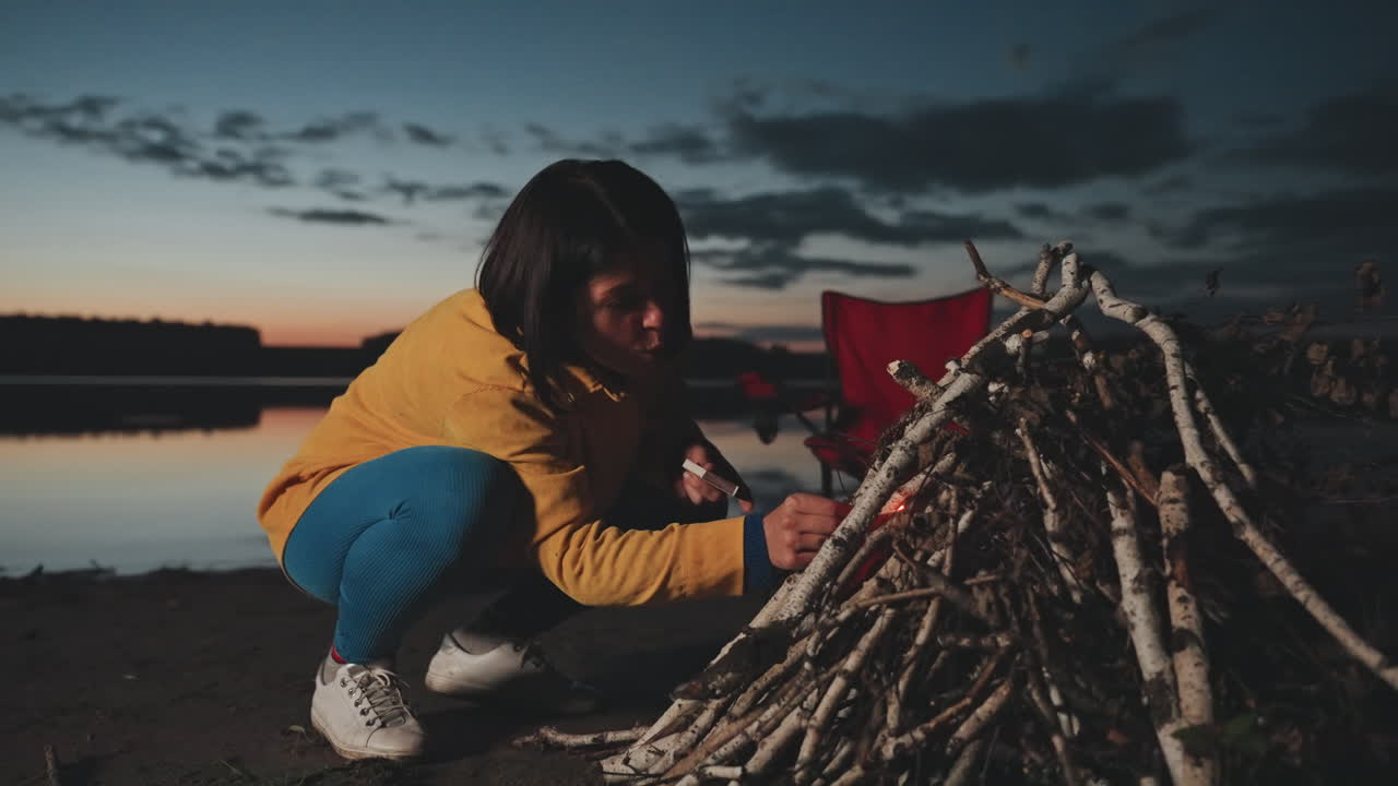 Aftencamp opsætning ved sø, camper arrangerer pinde-tipi og tændmateriale, metodisk, hands-on forberedelse til bål, rød stol og rolig vandbaggrund, praktisk overlevelsesfærdighed i stemningsfuldt tusmørke.