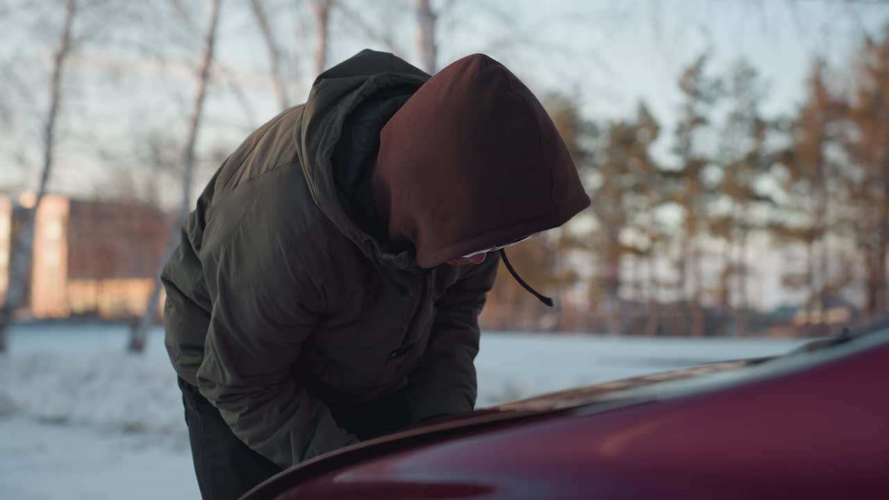 Man in hoodie bends over red car, struggling to open bonnet on snowy day with blurred view of residential buildings and bare trees in winter background