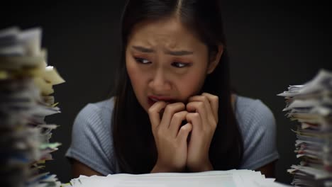 A young woman appears anxious and distressed while surrounded by towering piles of paperwork in a dimly lit office late at night, highlighting the stress of overwhelming tasks.