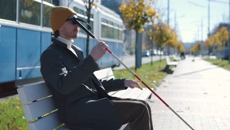 young blind man with smartphone sitting on bench in park in city, calling