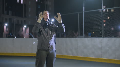 footballer in gloves tossing a soccer ball into the air and catching it during nighttime practice, urban backdrop with blurred building, bokeh light effects, and three people walking behind the fence