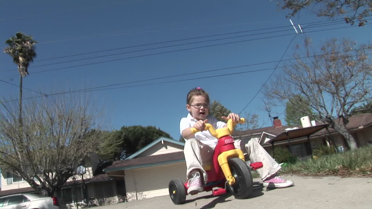 una niña pierde el equilibrio en un triciclo de plástico y rueda por un camino de entrada