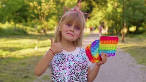 a little girl smiles and holds up a rainbow pop it toy in a park