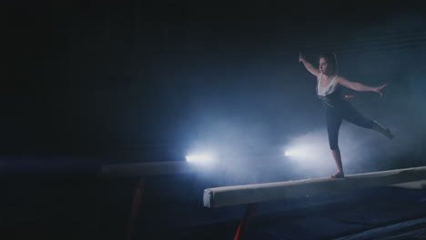 the girl performs a trick on a log in backlight and slow motion in sports gymnastic clothing. smoke and blue. jump and spin on the balance beam