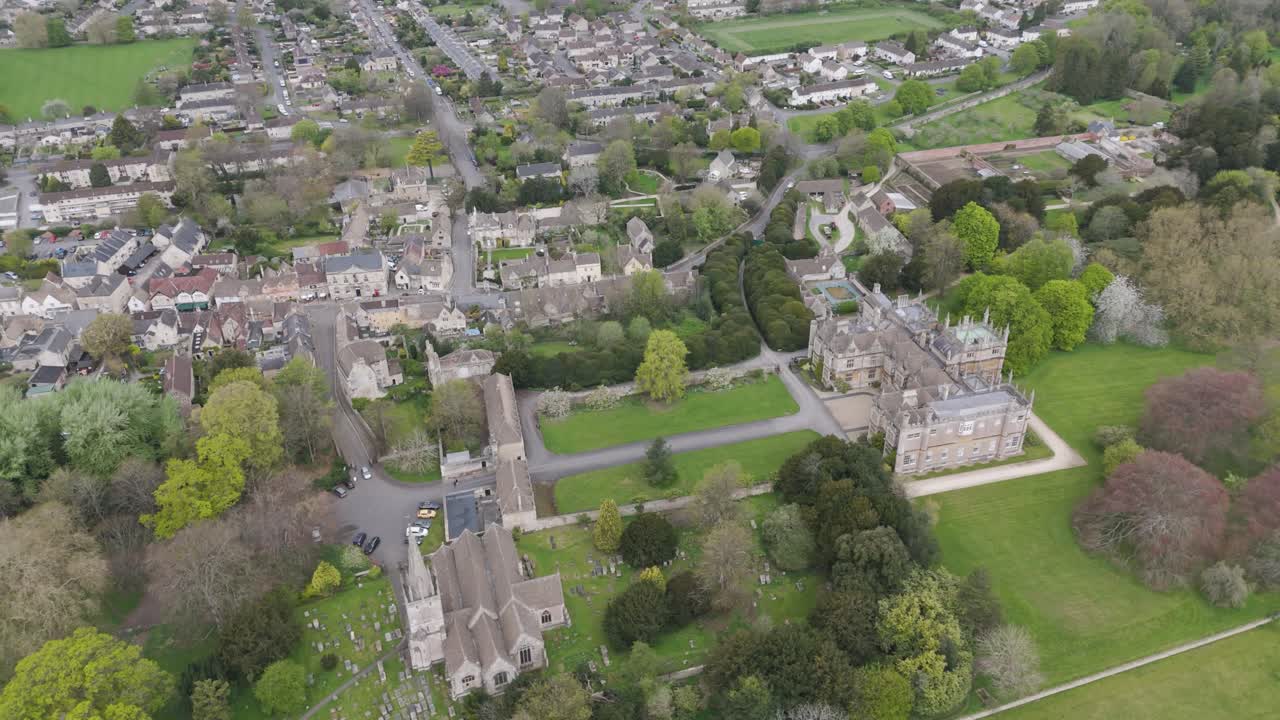 Wide aerial showing stately manor, formal gardens, tree-lined high street and patchwork countryside blending heritage architecture with small-town community life