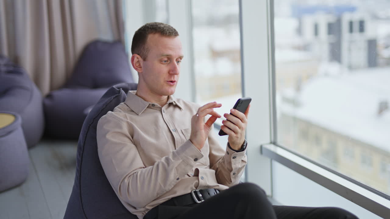 Video chat conversation at smartphone. Middle-aged man talks and smiles and waves his hand sitting in chair near window. Blurred backdrop.