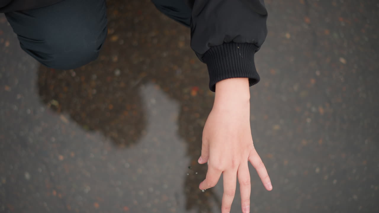 きらめく反射する池のそばにひざまずく女性。嵐の中、明るい水面にそっと触れる孤独な女性。雨の日に静かに輝く水たまりにひざまずく一人の女性。