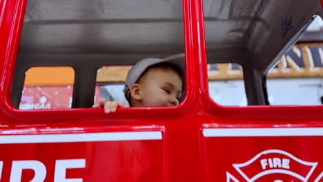 adorable baby boy face peeping from the red toy car he's sitting in. toddler pays inside the fire machine.