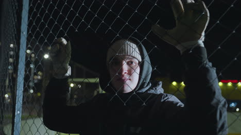 athlete standing behind chain-link fence during nighttime training session, wearing gloves and a hooded jacket, illuminated by urban lights