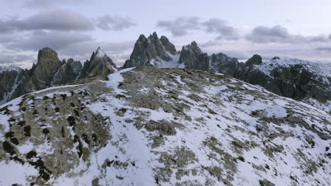 Aerial-view-over-a-snowy-ridge-in-front-of-the-Towers-of-Mordor,-winter-sunset-in-Dolomites,-Italy