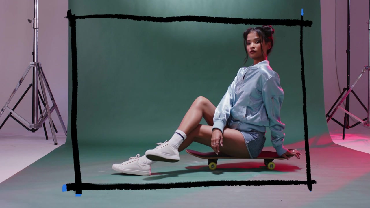 Camera cueing female model on skateboard, adjusting poses while black rectangle framing for fashion