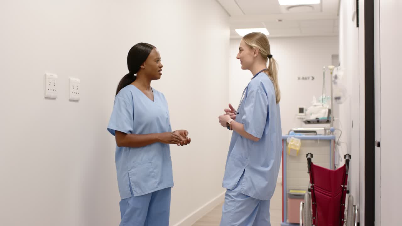 Female nurses talking in hospital corridor after shared joke, gesturing near red wheelchair