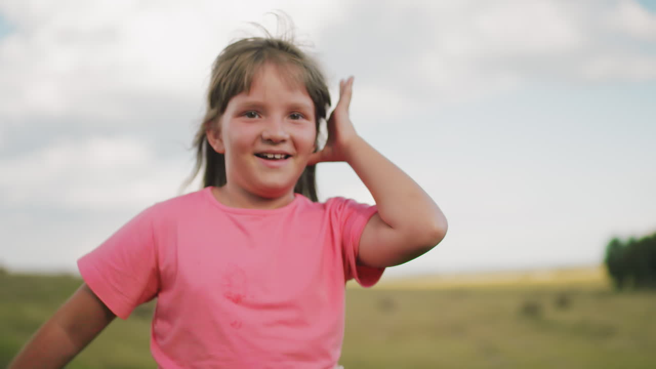 una joven vestida de rosa corre alegremente con el cabello soplado por el viento, sonriendo brillantemente contra una vasta tierra de cultivo borrosa, su movimiento despreocupado y expresión feliz