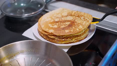 a person flips golden pancakes on a stovetop with a stack of fresh pancakes on a plate nearby