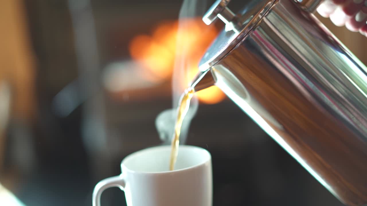 movimiento lento vertiendo café de prensa francesa caliente y humeante en una taza frente a una chimenea brillante