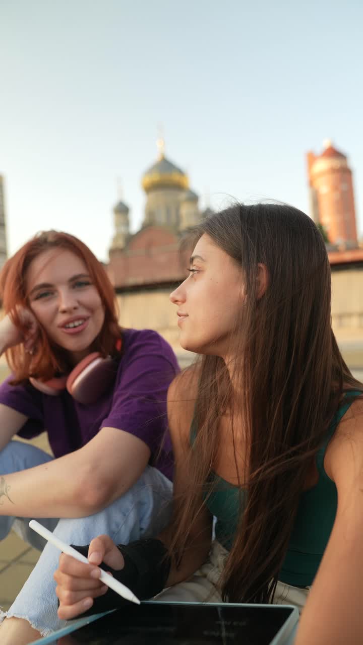 Due giovani amiche sedute su un tetto, che conversano mentre lavorano su un tablet