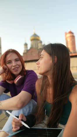 two young women friends sitting on a rooftop, having a conversation while working on a digital tablet.