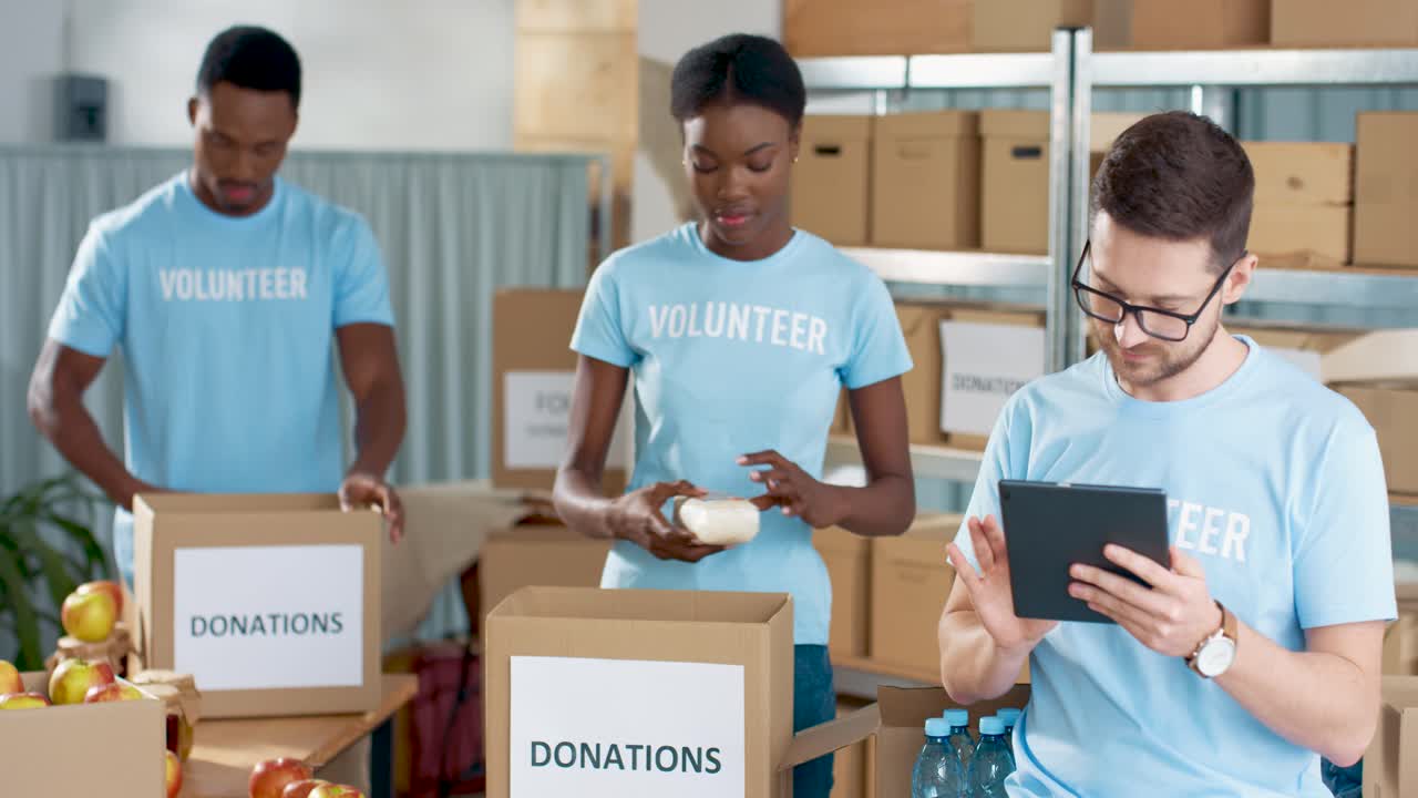 grupo multiétnico de voluntarios empacando cajas con comida y ropa y escribiendo en una tableta en un almacén de caridad