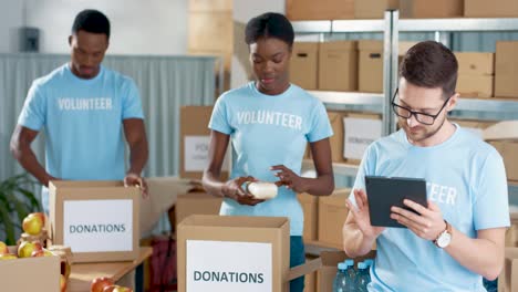 Multiethnic-group-of-volunteers-packing-boxes-with-food-and-clothes-and-typing-on-tablet-in-charity-warehouse
