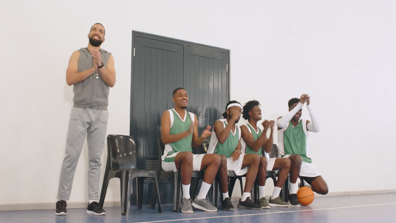 Männliche Basketballspieler verschiedener Ethnien und ihr Trainer, der das Spiel von Stühlen aus in der Sporthalle begleitet.