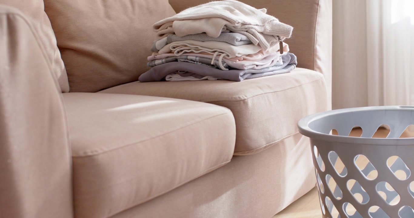 Camera is panning beige sofa with folded laundry near window as daylight is shifting conveying calm