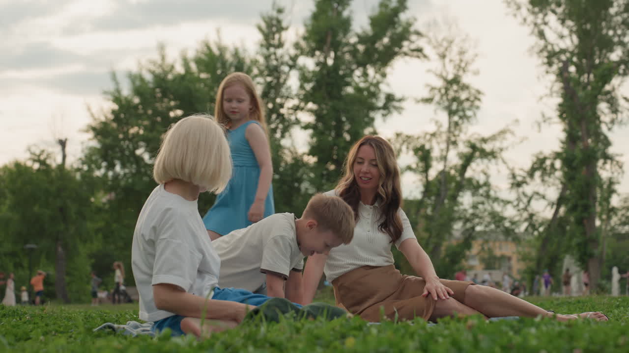 young mothers sitting on mat in park calling running kids to join, laughter and playful energy, summer grass backdrop, relaxed family moment, children approaching, warm light