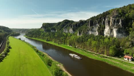 Aerial-Drone-View-Of-River-Cruise-Boat-Gently-Sailing-Along-Elbe-River-Past-Picturesque-Landscape-Of-Rathen,-Germany