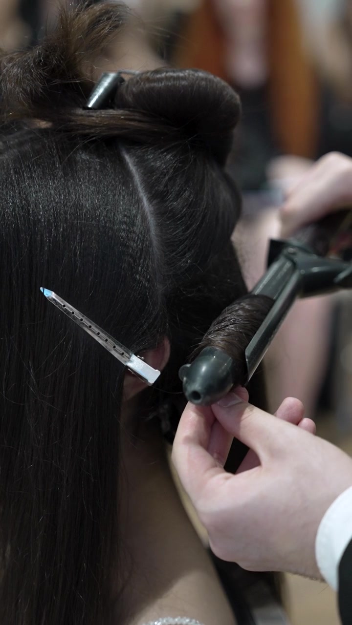 Un parrucchiere sta arricciando capelli lunghi in un salone. Lo stilista tiene un ferro arricciacapelli mentre la cliente è seduta su una sedia. Questo accade nel pomeriggio con altri clienti presenti