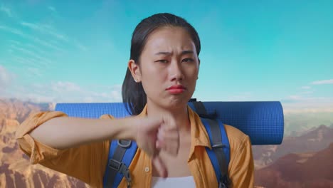 close up of asian female hiker with mountaineering backpack showing thumbs down gesture while traveling at the top of mountain
