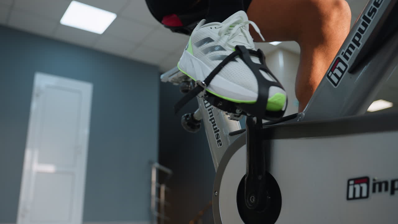 Lower angle view of man legs pedaling on impulse stationary bike in gym, showing branded frame, foot strap, and tile floor under bright ceiling lights with blurred gym interior background
