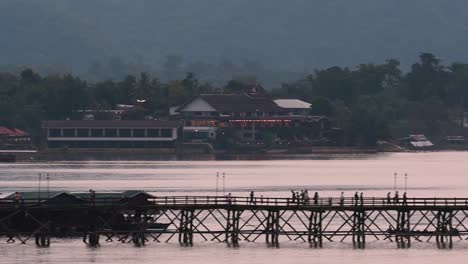 Mon-Bridge-and-a-longboat-followed-speeding-behind-the-structure-while-people-are-walking-on-the-bridge,-silhouetting-as-it-was-getting-dark,-in-slow-motion