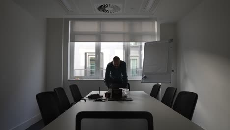 An-Asian-office-employee-standing-crouched-over-reaching-down-as-he-works-on-a-laptop-computer,-the-man-alone-in-an-empty-meeting-room-making-last-minute-changes-to-a-presentation