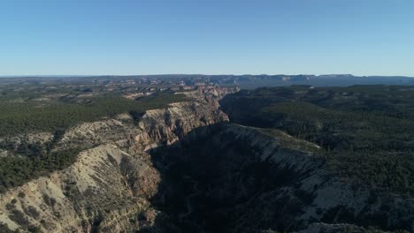 Drone-aerial-flyover-of-river-valley-canyon-in-Utah-with-green-trees,-bushes,-sandy-cliffs,-hills-and-canyons-in-the-distance-under-a-blue-sky