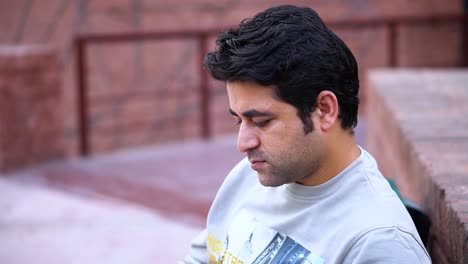 Close-up-shot-of-a-South-Asian-man-looking-down-while-sitting-outside-on-a-bench-reading-a-book