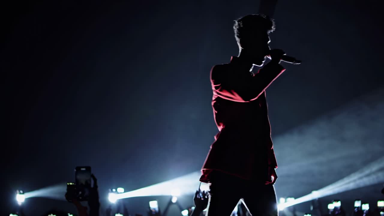 Young male singer performing live on a dark stage, wearing a red jacket, illuminated by spotlights and fans' smartphones recording the show