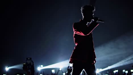 young male singer performing live on a dark stage, wearing a red jacket, illuminated by spotlights and fans' smartphones recording the show