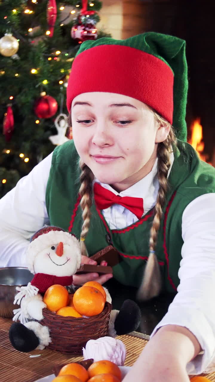 Girl in elf costume sitting at festive table. Funny elf takes many chocolate pieces from plate in Christmas decorated room. Vertical video