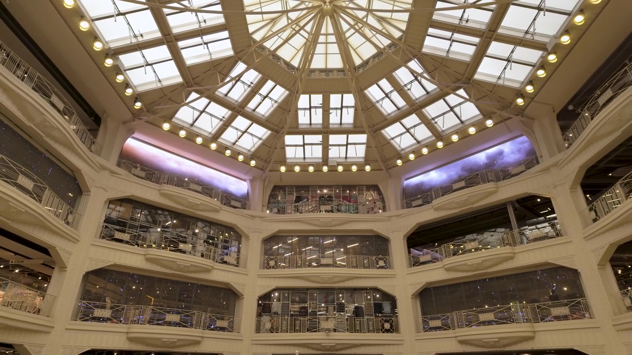 Una ripresa dal basso di un lussuoso centro commerciale a più livelli, caratterizzato da un grande atrio in vetro e un'elegante architettura bianca. Adatto per contenuti di viaggio, vendita al dettaglio e stile di vita urbano.