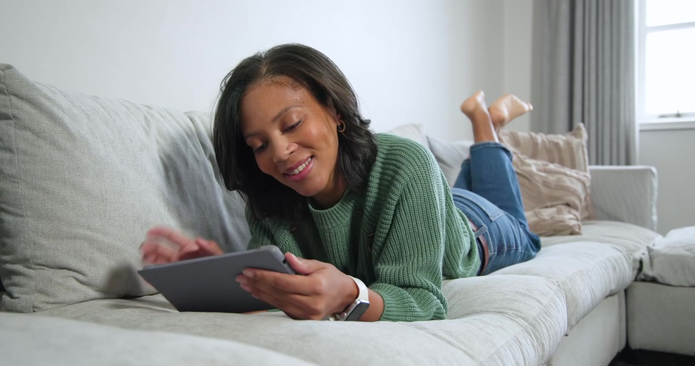African American woman lying on sofa holding gray tablet in living room, watching clip, smiling