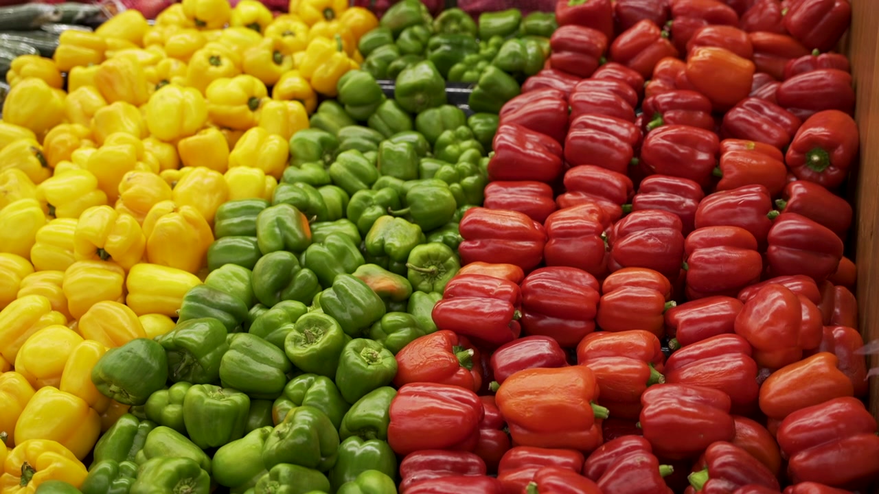 File di peperoni gialli, verdi e rossi formano vivaci bande diagonali nella sezione ortofrutta di un supermercato.