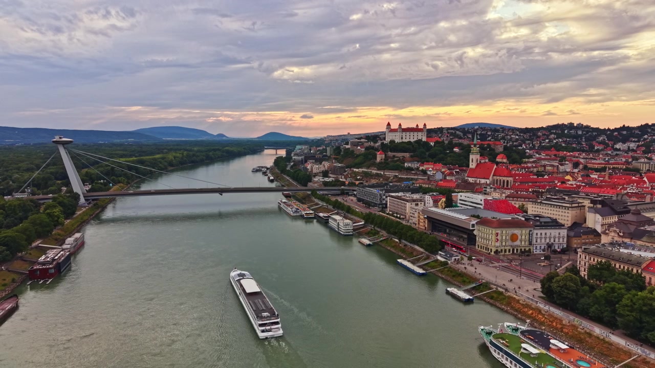 Byudsigt over Bratislava med en bro, flod og bygninger under en overskyet himmel ved solnedgang